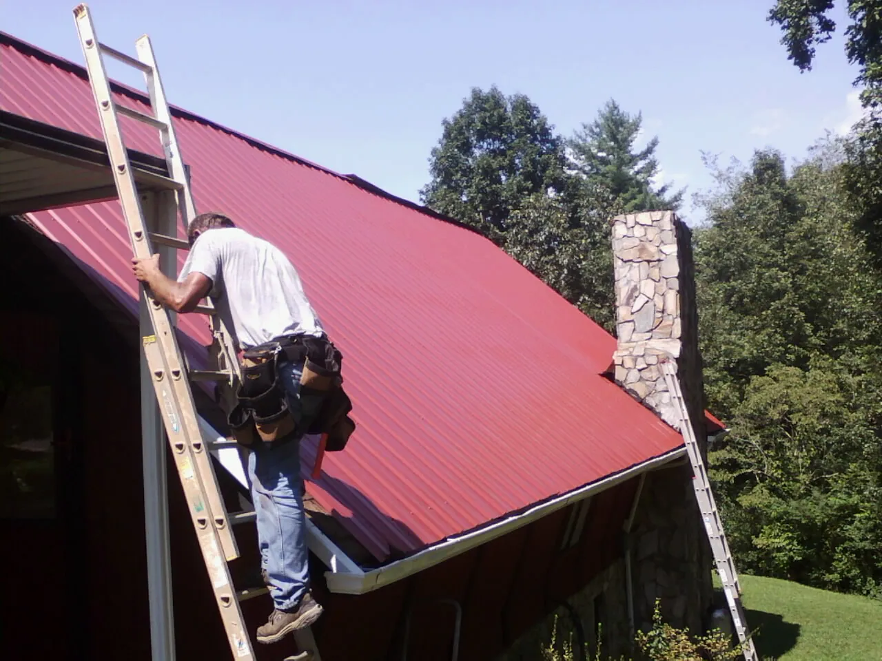 Roofer on a ladder beside a red metal roof and stone chimney — Soffit & Fascia Repair by Roofing Brothers in Lumpkin